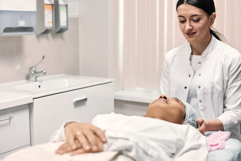 A clinician leans in to talk with a client resting on a treatment bed before a procedure